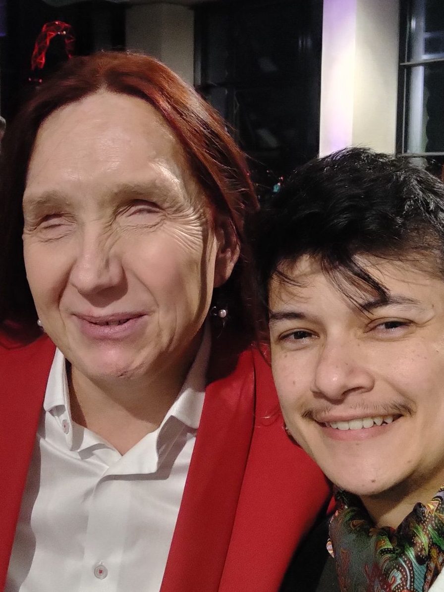 Sarah and Pk sitting beside each other at an awards ceremony, smiling at the camera. Sarah is a white woman with red hair, and is wearing a red suit with a white shirt. Pk has fair brown skin, black hair and is wearing a cravat and a black tuxedo jacket, with a white shirt.